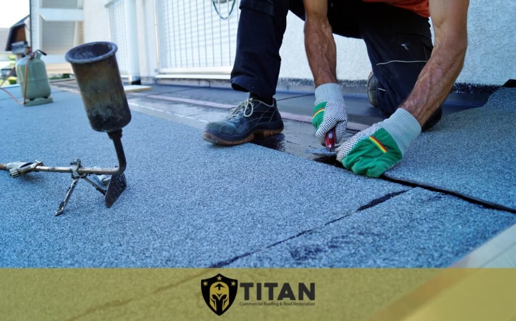 A worker in gloves uses a cutting tool to install roofing material on a flat roof. A torch and the Titan logo are visible in the foreground.