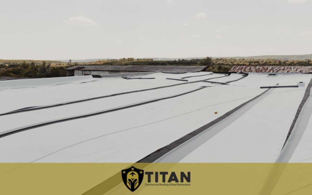 Flat commercial rooftop with white membrane material and visible seams, set in a rural area; Titan Commercial Roofing & Roof Restoration logo and branding overlay at the bottom.