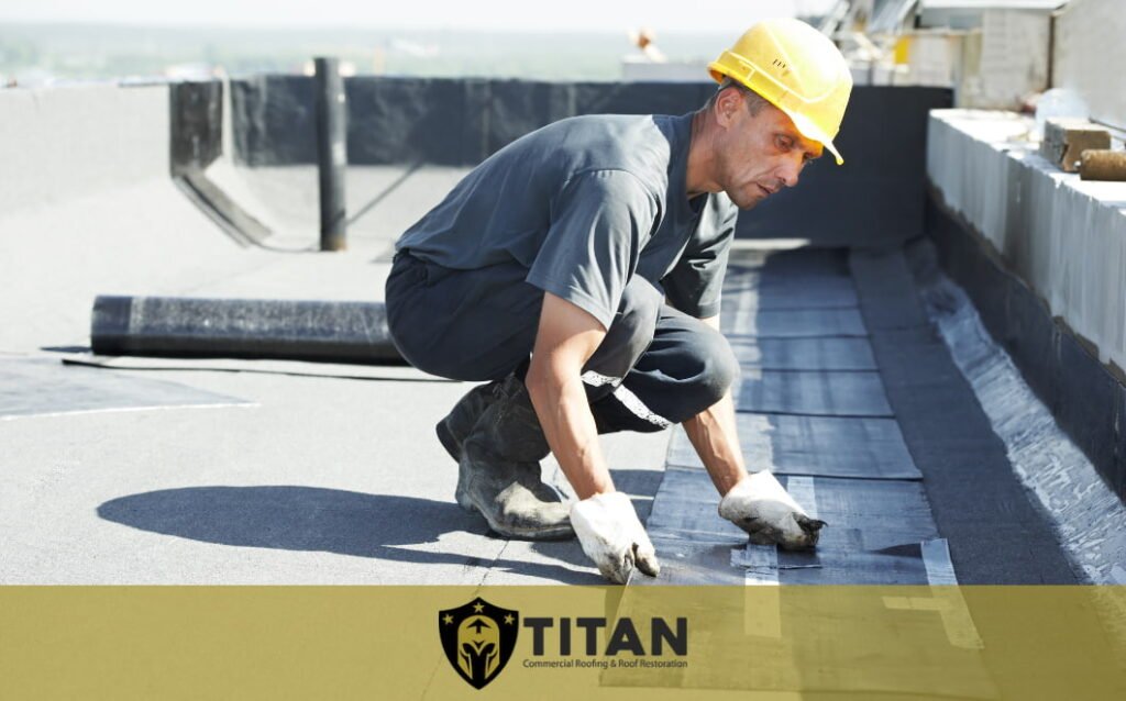 A worker in a yellow hard hat installs roofing material on a flat roof. The logo and text "Titan Commercial Roofing & Roof Restoration" appear at the bottom.
