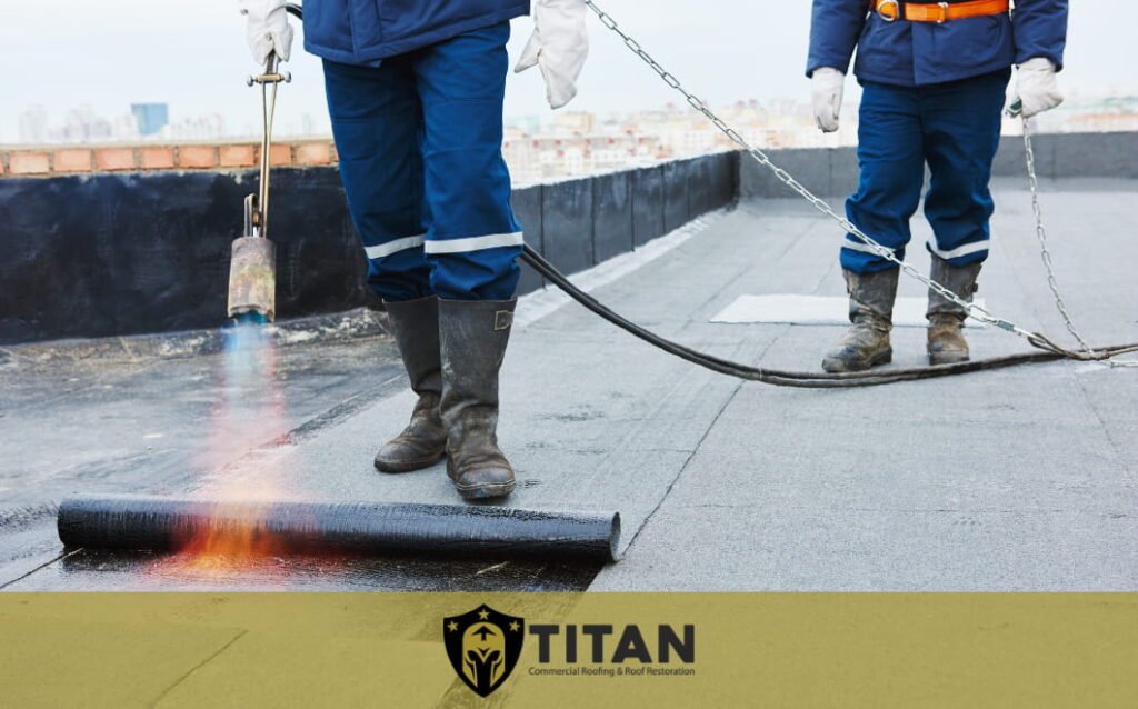 Two workers in safety gear apply heat to roll roofing material on a flat commercial roof, with the Titan company logo visible at the bottom.